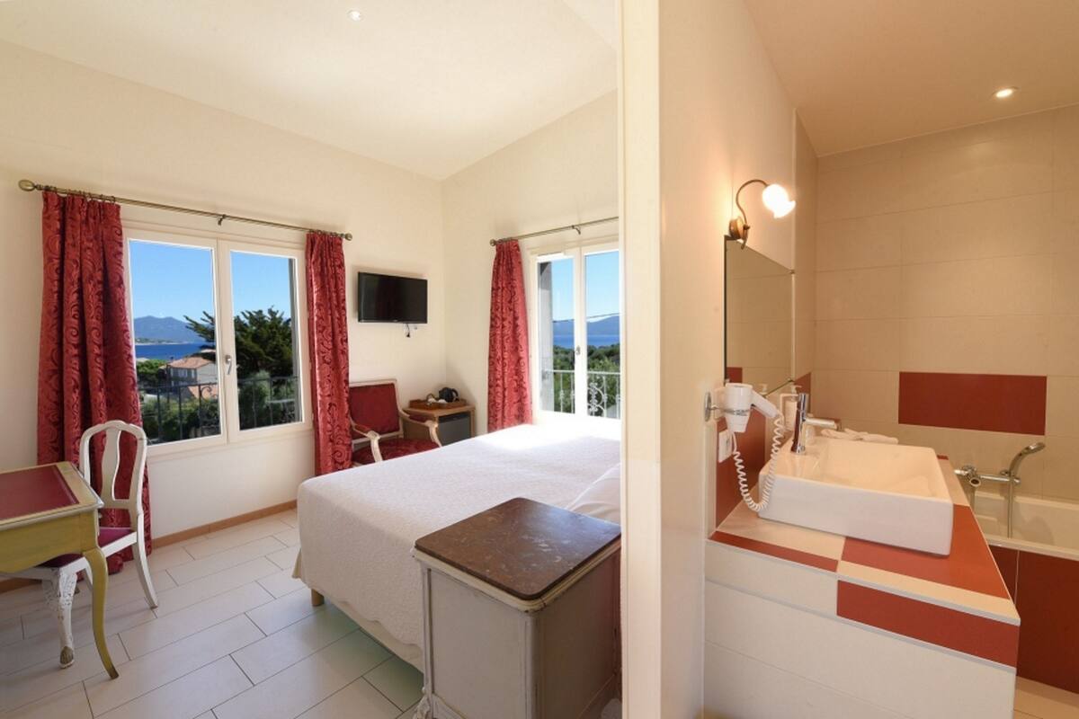 A hotel room with a bed, desk, chair, and a bathroom with a sink and mirror.