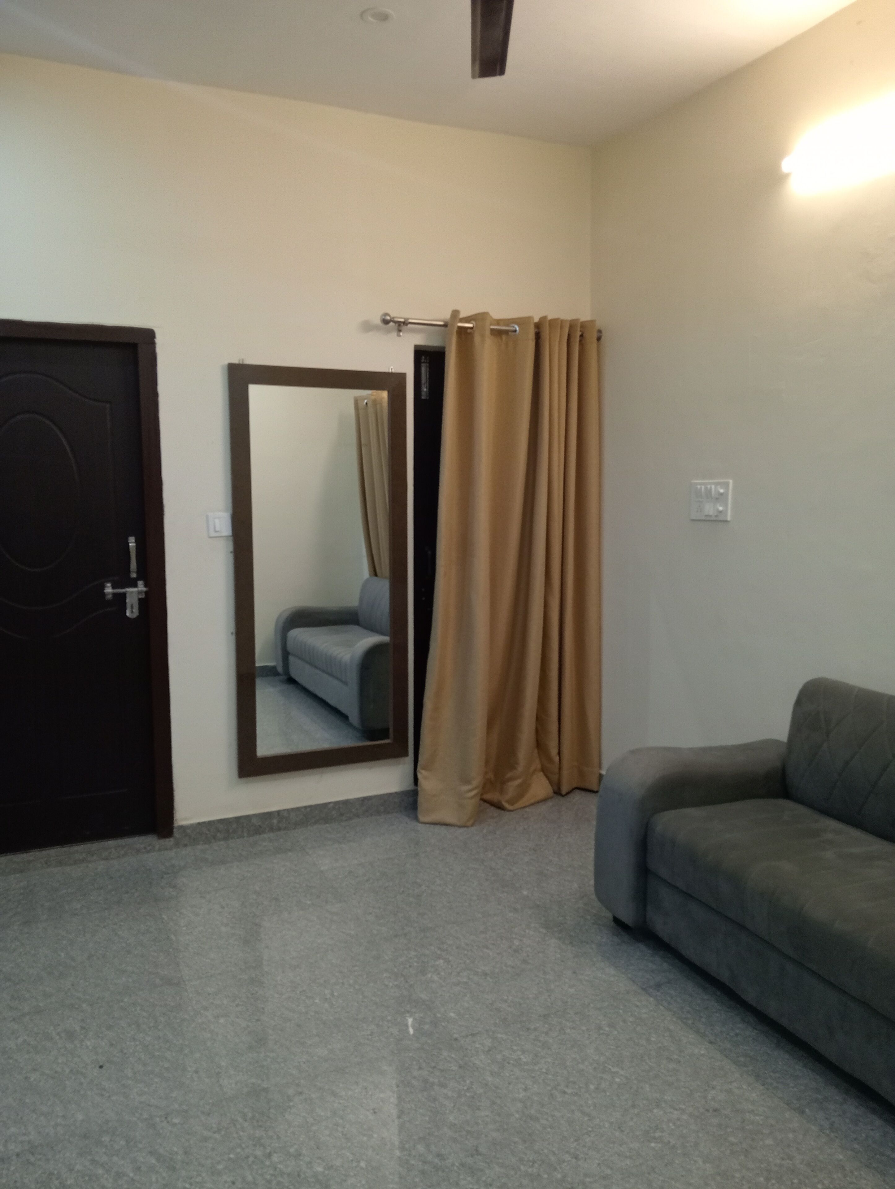 Una habitación con un sofá gris, una puerta, un espejo y una cortina.