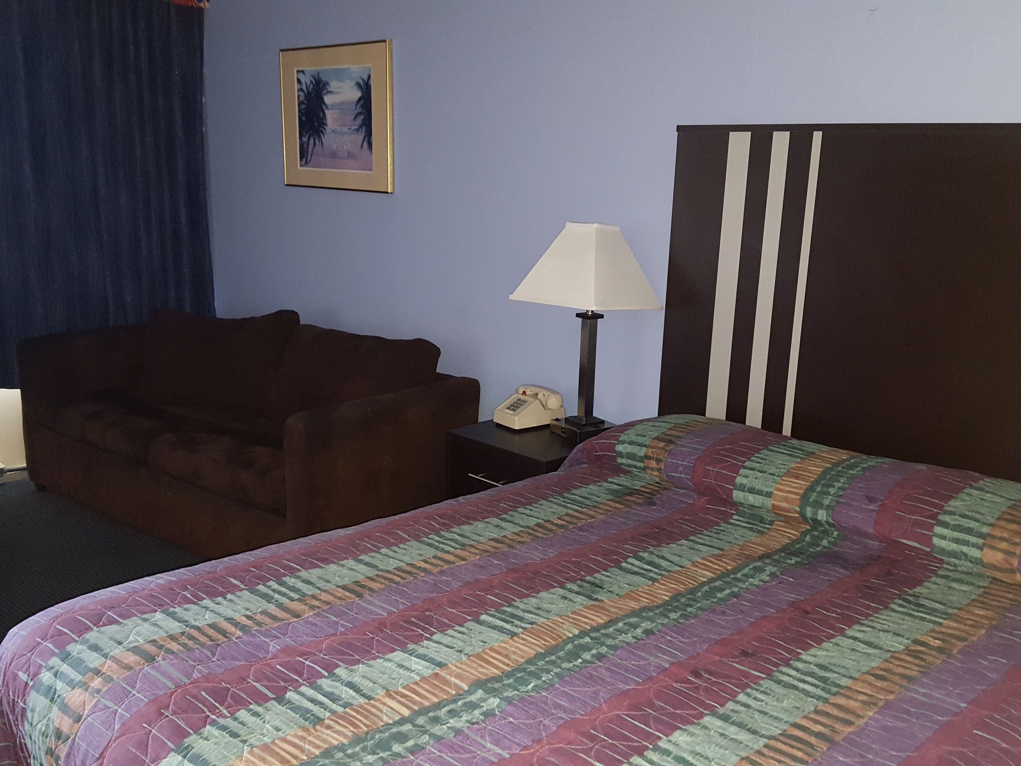 A hotel room with a bed, a brown armchair, a nightstand with a lamp, and a painting on the wall.
