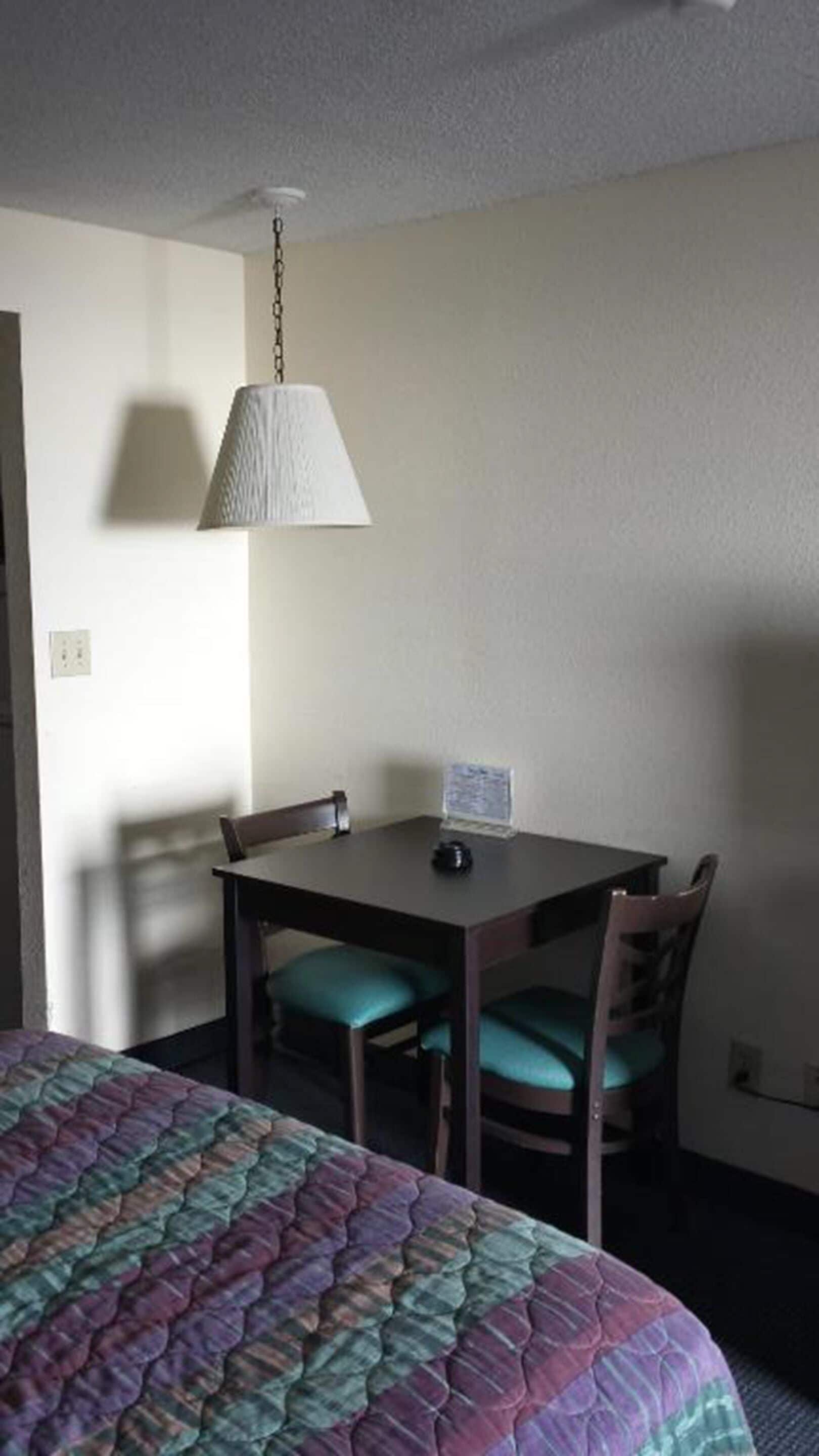 A hotel room with a bed, a table with chairs, a hanging lamp, and a small table with a lamp.