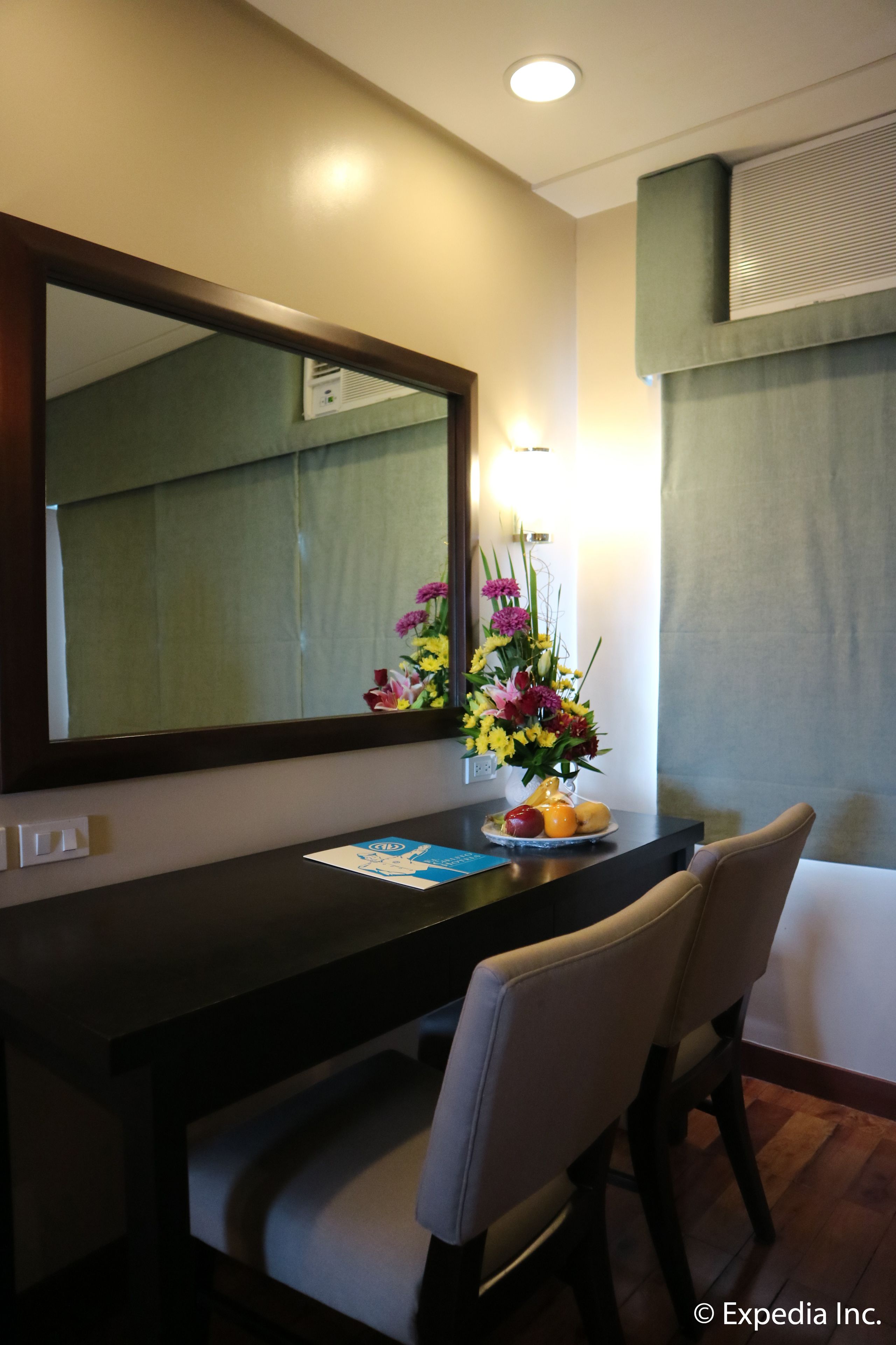 Habitació d'hotel amb escriptori de fusta fosca, un mirall, una gerra amb flors i una cadira.