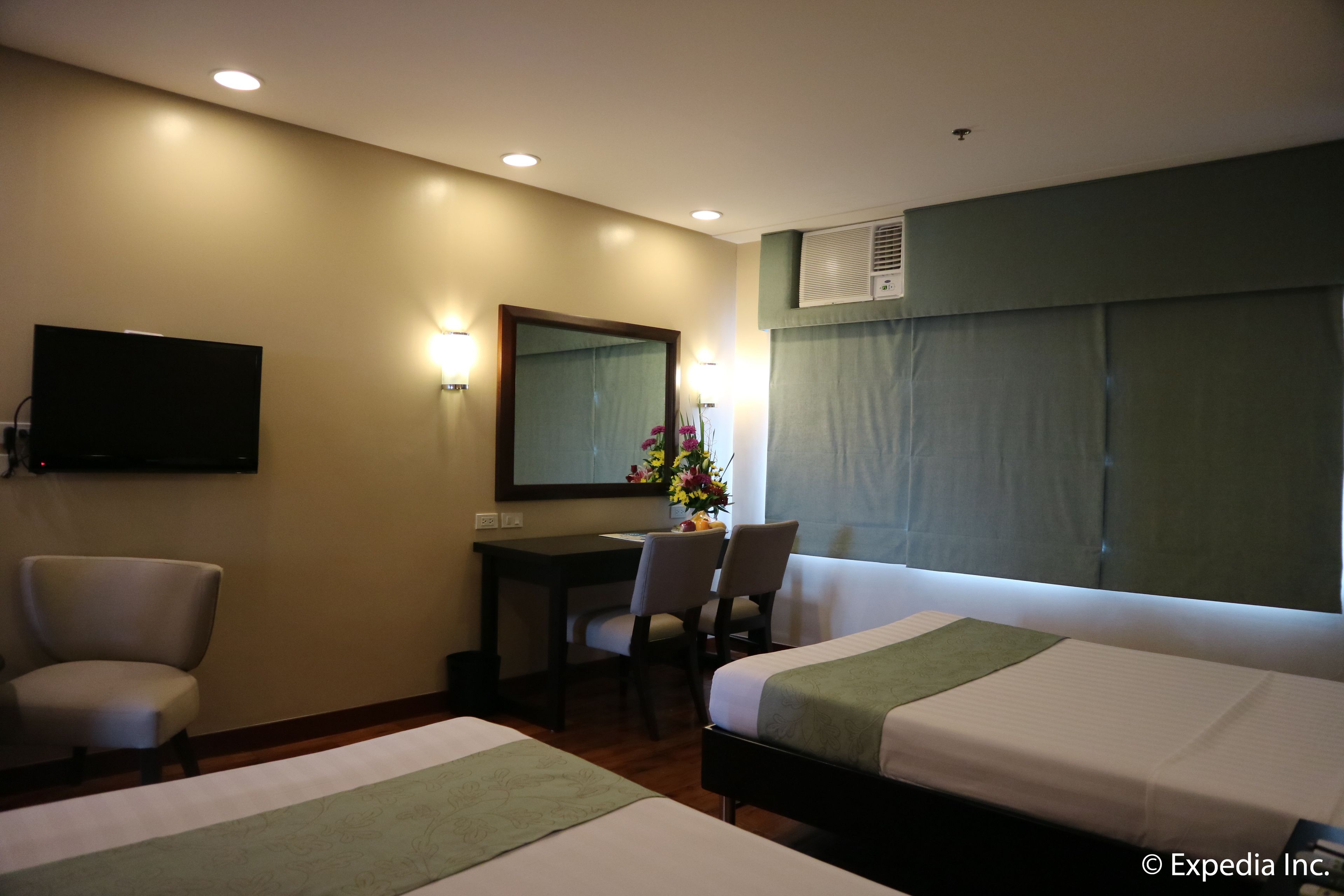 Quarto de hotel com duas camas, uma escrivaninha, uma cadeira, uma televisão e um vaso de flores.