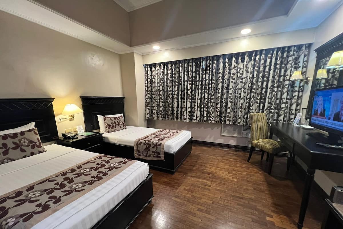 A hotel room with two beds, a desk with a television, a chair, and a window with patterned curtains.