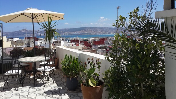 A rooftop terrace with a table and chairs, potted plants, and a view of the sea and distant hills. - Riad Arous Chamel (Tangier)