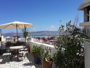 Terraço no telhado com mesa e cadeiras, plantas em vasos e vista para o mar e colinas ao longe.