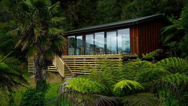 A modern wooden cabin with large windows, surrounded by lush greenery and ferns. - The Lodge At Te Rawa - Boat Access Only (Te Rawa)