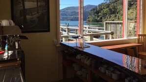 A kitchen with a coffee machine, a window with a view of a dock and mountains, and shelves with kitchenware. - The Lodge At Te Rawa - Boat Access Only (Te Rawa)
