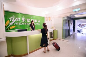 Un comptoir d’accueil d’hôtel aux couleurs vert et blanc, une enseigne indiquant « KIWI EXPRESS HOTEL », et une personne se tenant derrière le comptoir.
