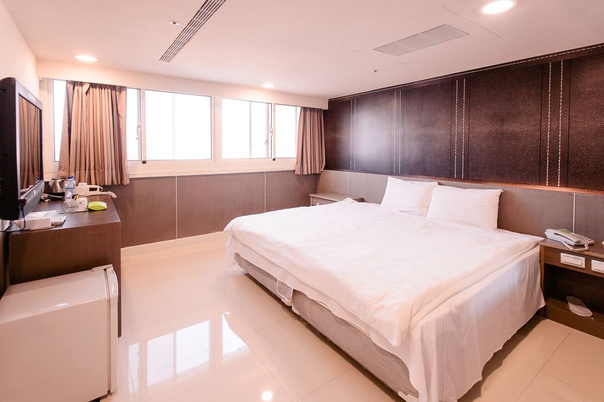 A hotel room with a large bed, a desk with a computer, a refrigerator, and a window with curtains.