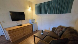 A living room with a flat-screen TV mounted on the wall, a wooden dresser, a white microwave, a brown sofa with patterned cushions, and a small table with a plant. - Shannon Motel (Fort Nelson)