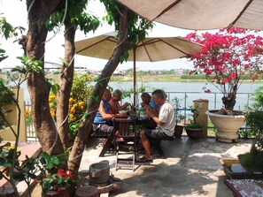 A group of people dining at an outdoor table under a canopy, surrounded by potted plants and a view of a body of water. - Homeland River Homestay (Da Nang)