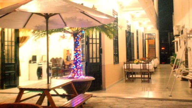 Un área de comedor al aire libre con una mesa y bancos bajo un paraguas grande, rodeada por un edificio con piso de baldosas y una palmera con luces de colores.