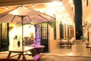 An outdoor dining area with a table and benches under a large umbrella, surrounded by a building with a tiled floor and a palm tree with colorful lights. - Homeland River Homestay (Da Nang)