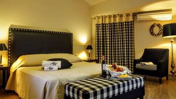 Habitación de hotel con una cama grande, un reposapiés a cuadros, una silla y una mesa con una botella y vasos.