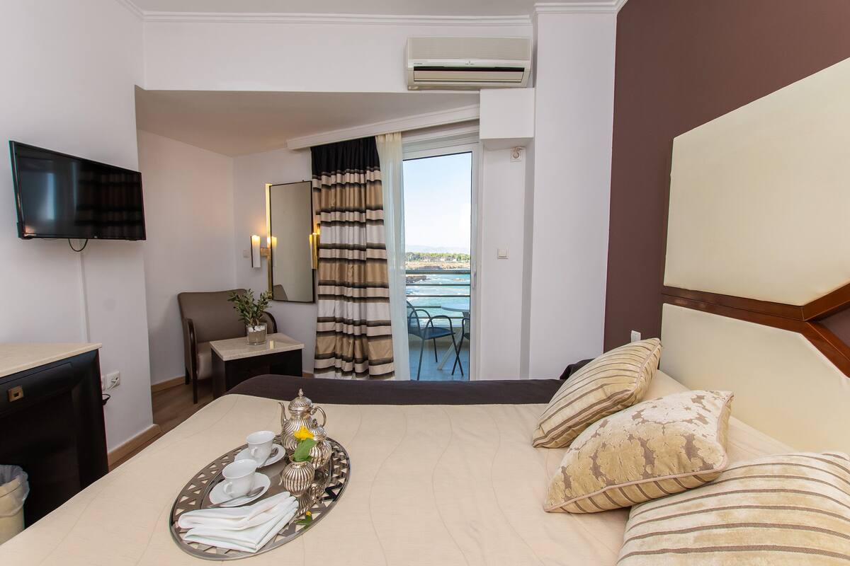 Ein Hotelzimmer mit Bett, Fernseher, Esstisch mit Teewagen und Tassen, Stuhl und Meerblick vom Balkon.