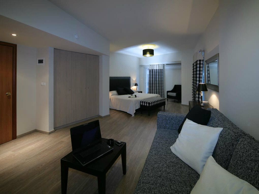 A modern hotel room with a sofa, a bed, a desk with a laptop, and a wardrobe.