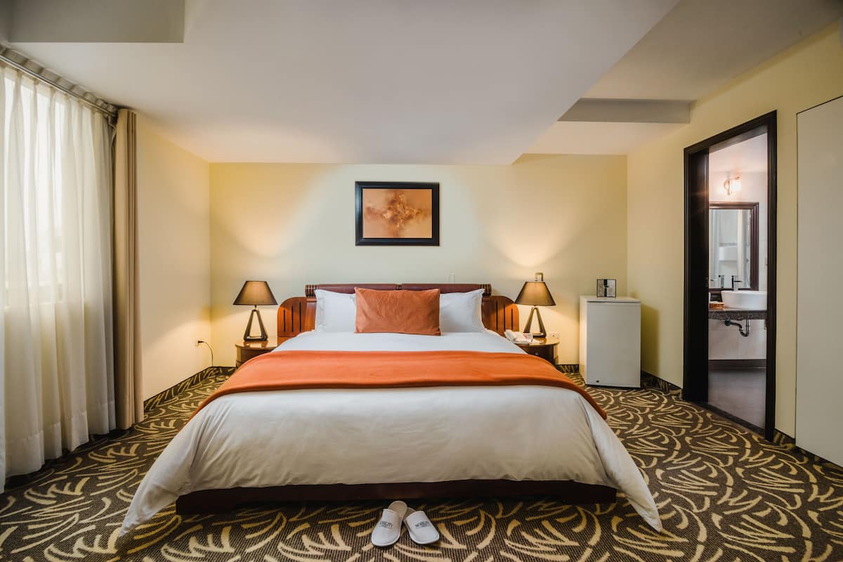 A hotel room with a large bed, two bedside tables with lamps, a small cabinet, and a door leading to a bathroom.