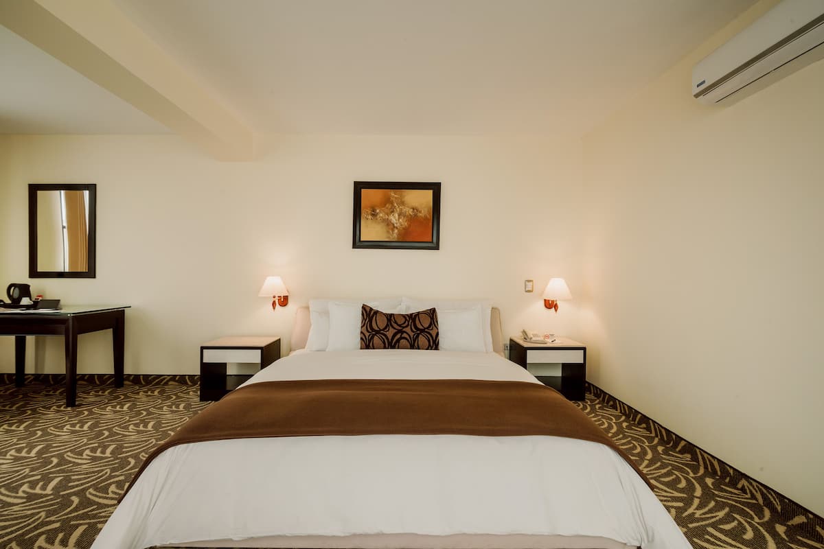A hotel room with a large bed, two bedside tables with lamps, a desk, a chair, a mirror, and a wall-mounted air conditioner.