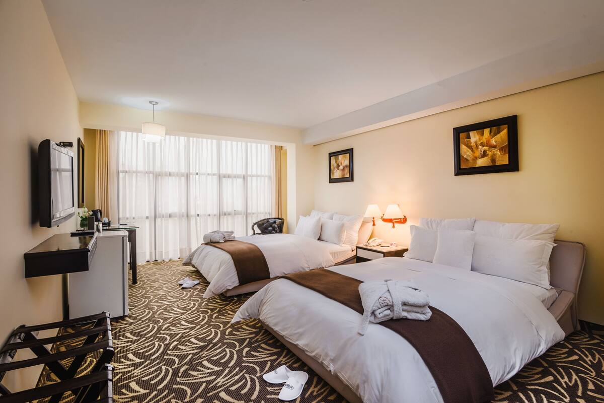 A hotel room with two beds, a desk, a chair, a TV, and a window with curtains.