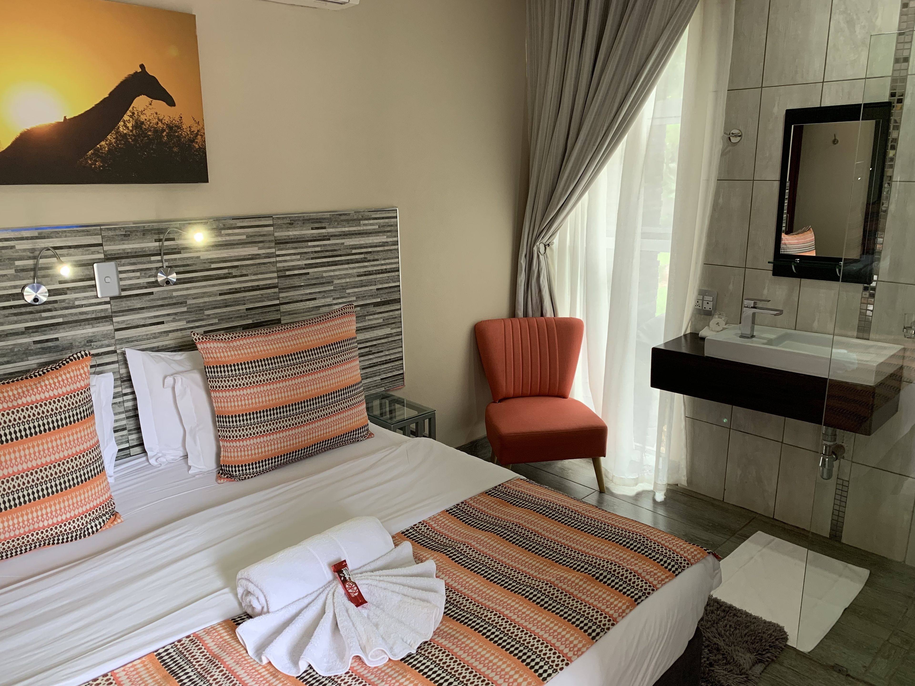 A hotel room with a bed, a chair, a bathroom with a sink and mirror, and a painting on the wall.