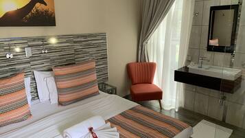 A hotel room with a bed, a chair, a bathroom with a sink and mirror, and a painting on the wall.