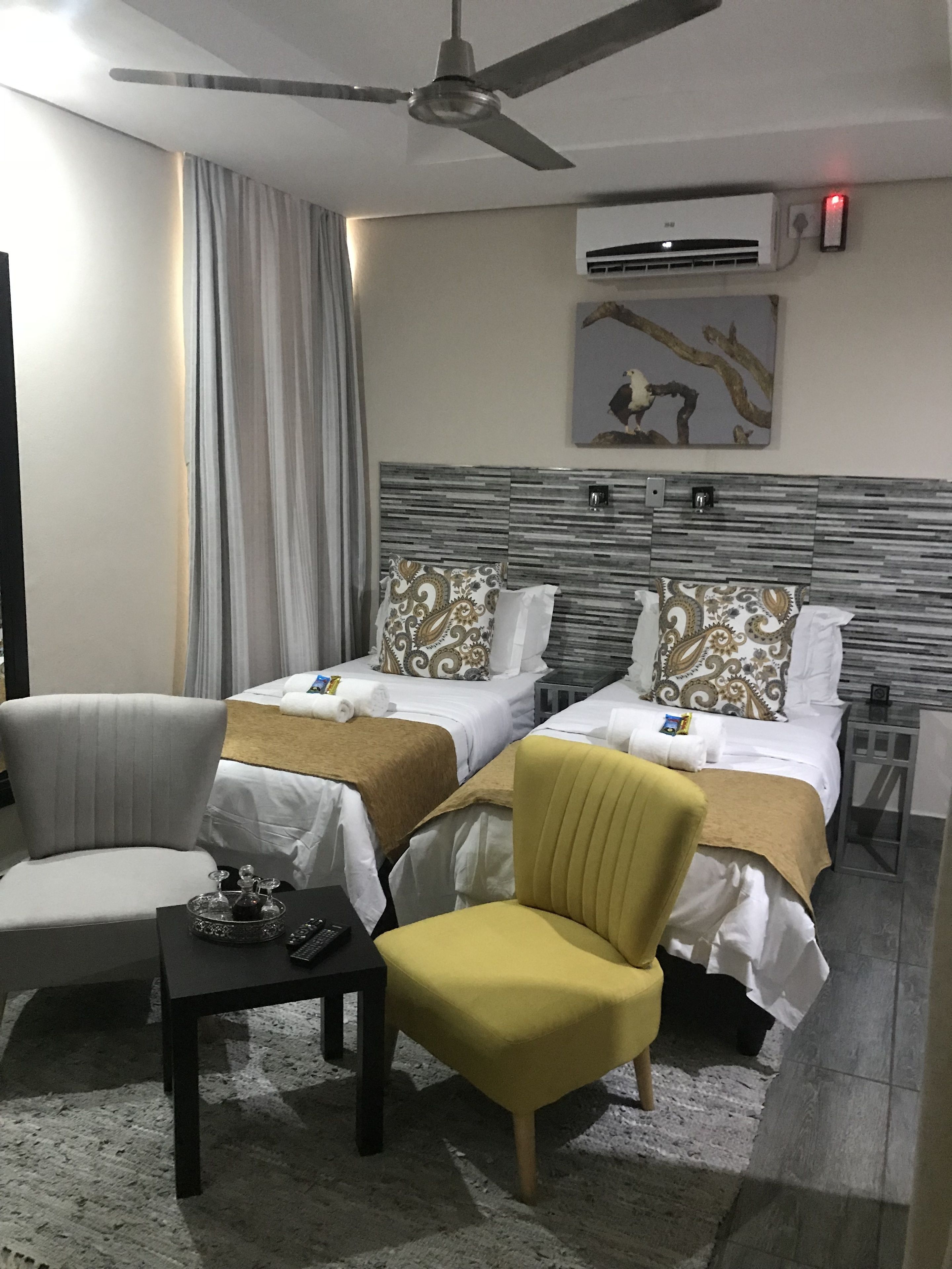 A hotel room with two beds, a chair, a small table, an air conditioning unit, and a ceiling fan.