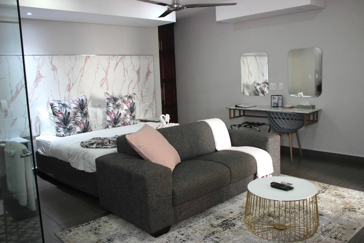 Un dormitorio moderno con una cama, un sofá gris, una mesita redonda y un espejo grande.
