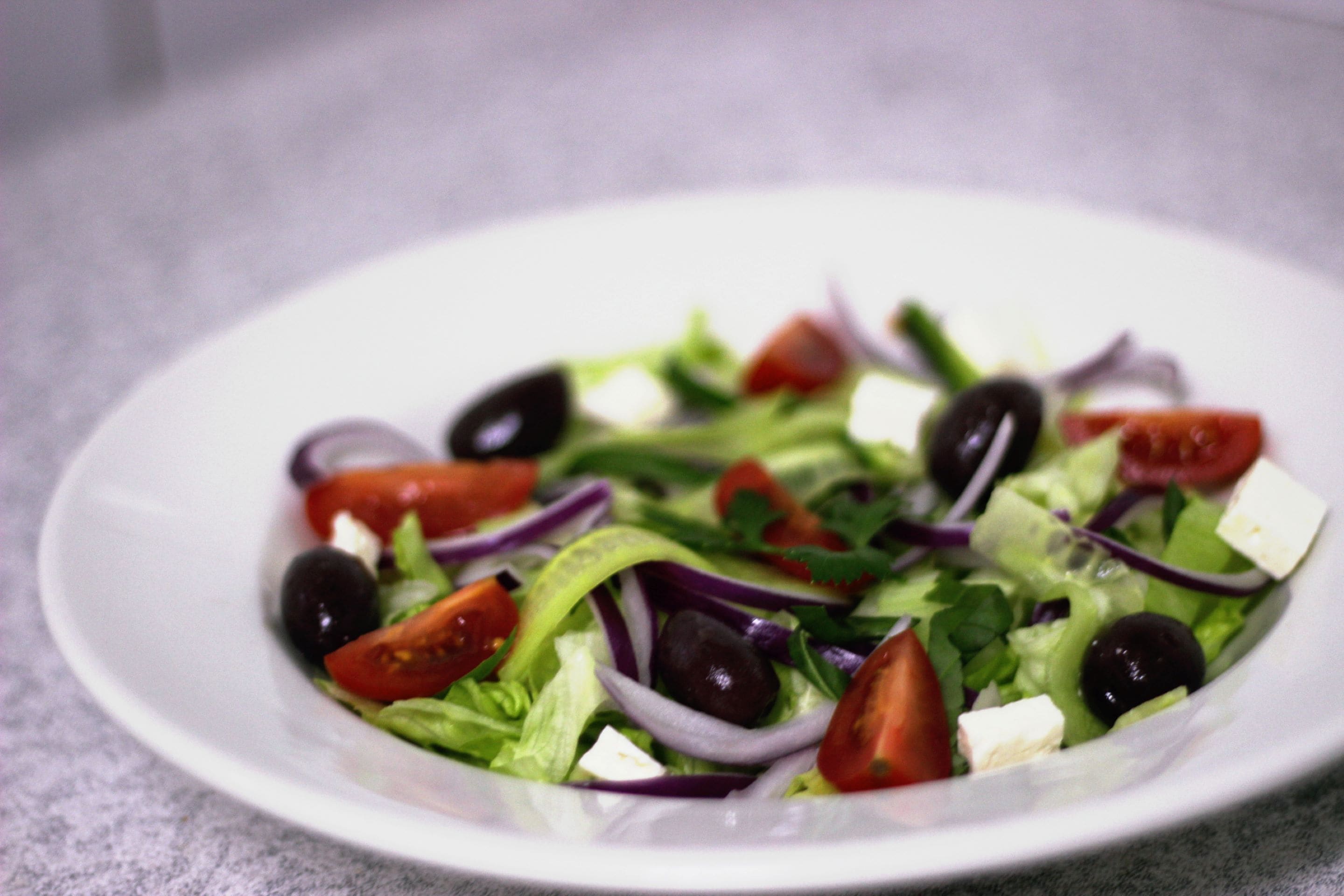 Une assiette blanche contenant une salade composée de laitue, de tomates, d’oignons rouges, d’olives noires et de fromage feta.