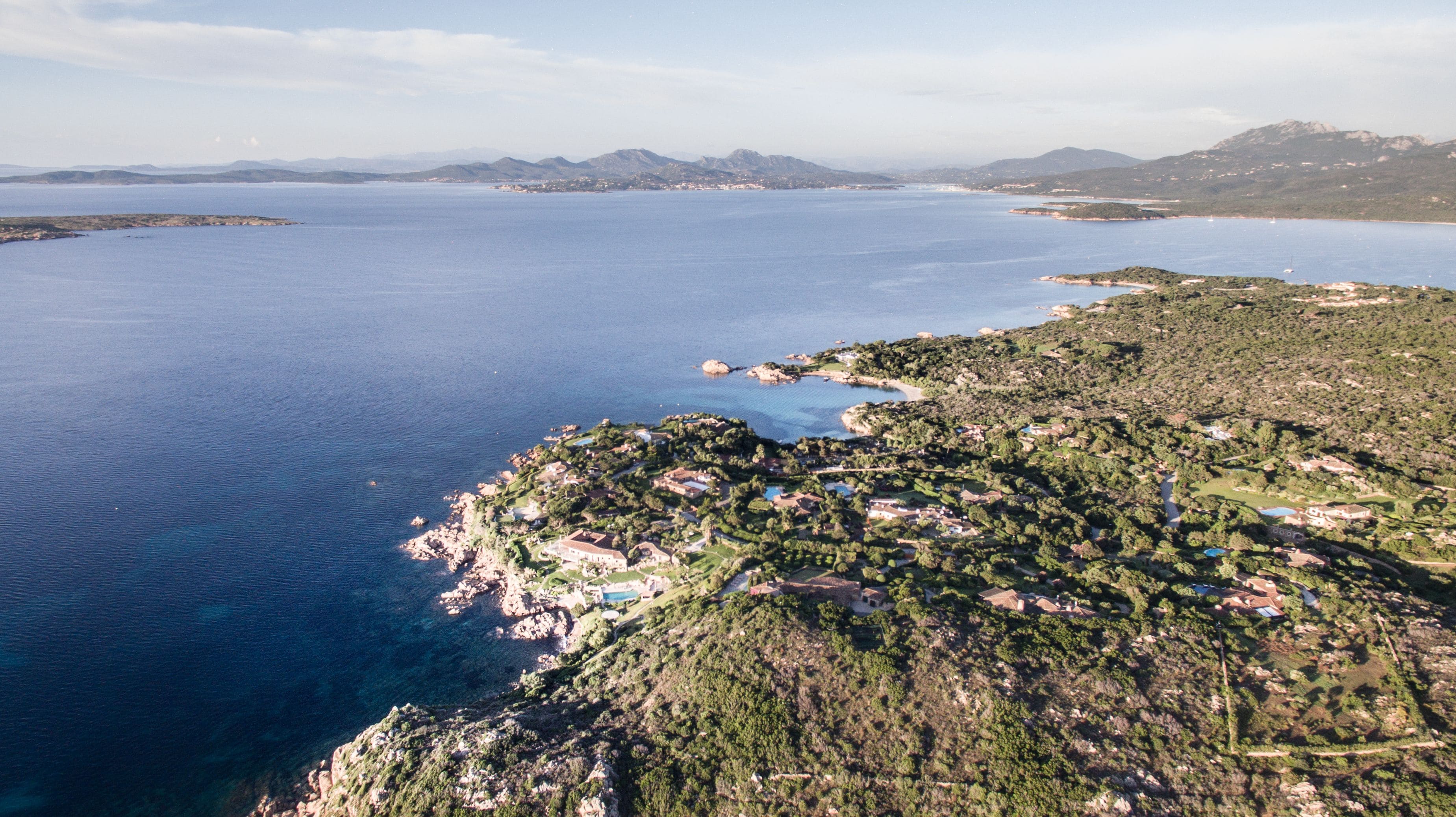 Vista aérea de uma área costeira com uma pequena cidade, mar azul cristalino e montanhas ao fundo.