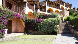 A traditional stone building with multiple balconies and arches, surrounded by lush greenery and purple flowers. - Residence Costa Ruja (Olbia)