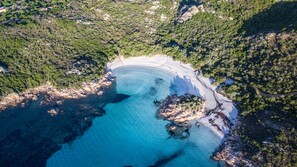 An aerial view of a coastal area with a sandy beach, clear blue waters, and surrounding forested hills. - Residence Costa Ruja (Olbia)