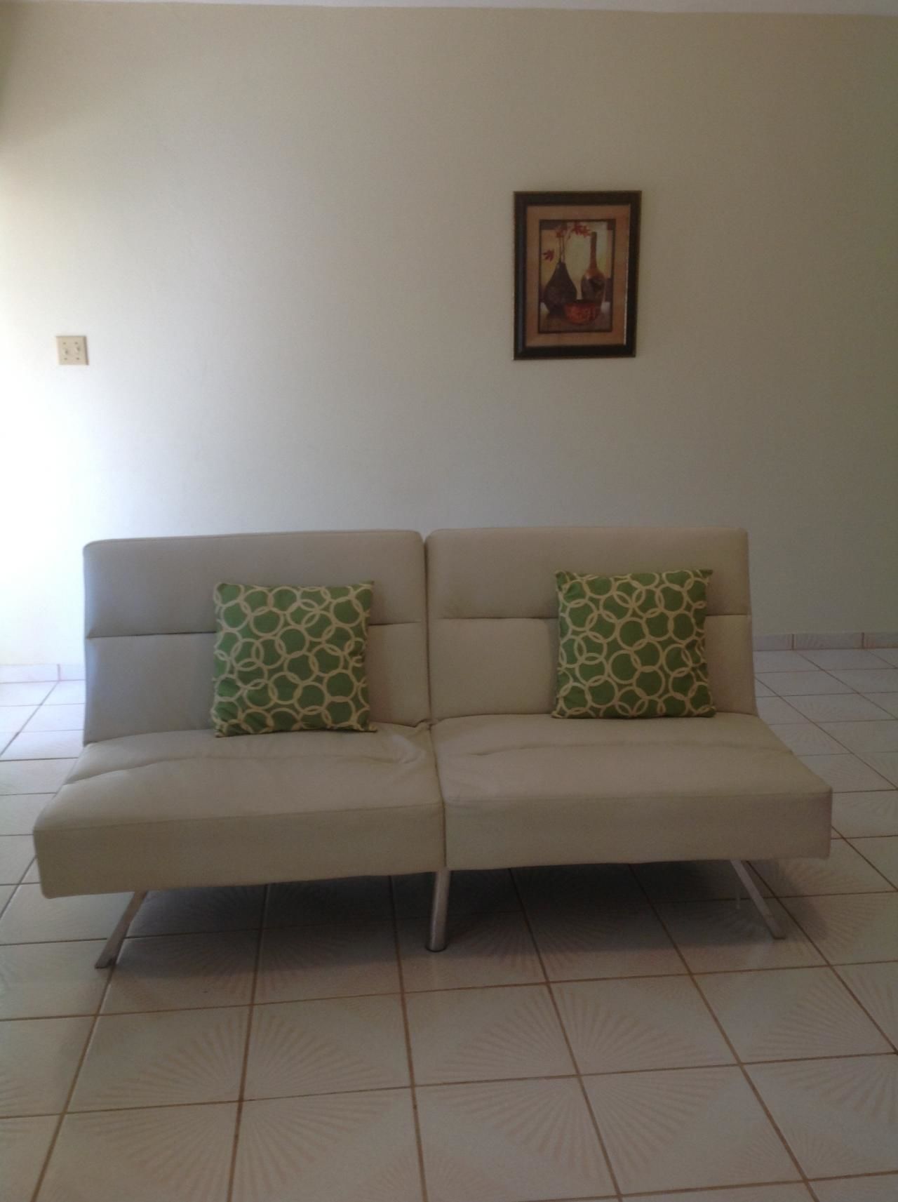 Un sofá color beige con cojines estampados verdes en una habitación con suelo de baldosas y un cuadro en la pared.
