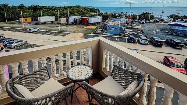 A balcony with wicker chairs and a table, overlooking a parking area and a marina with boats.