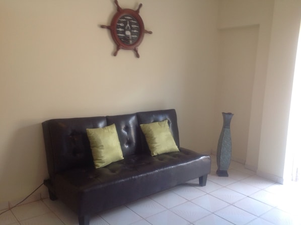 Un sofá de cuero oscuro con dos cojines verdes frente a una pared lisa, un reloj de babor de madera y un jarrón alto.