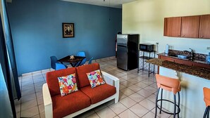 A living room with a red sofa, a ceiling fan, a kitchen with a refrigerator, and a blue wall.