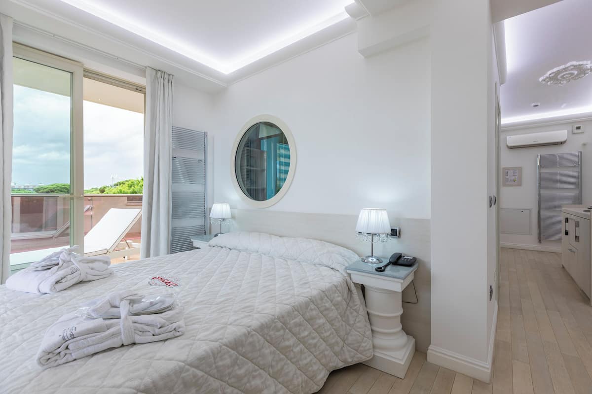 A hotel room with a bed, a round mirror, a bedside table with a lamp, and a view of the outdoors.