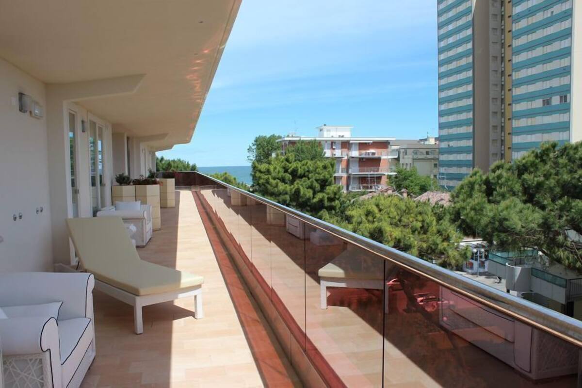 A balcony with sun loungers and a glass railing overlooking a coastal area with buildings and trees.