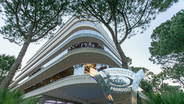 A multi-story hotel with a curved facade, a prominent sign for the Paradiso Hotel, and lush greenery surrounding the building. - Paradiso Hotel Bovelacci (Cervia)