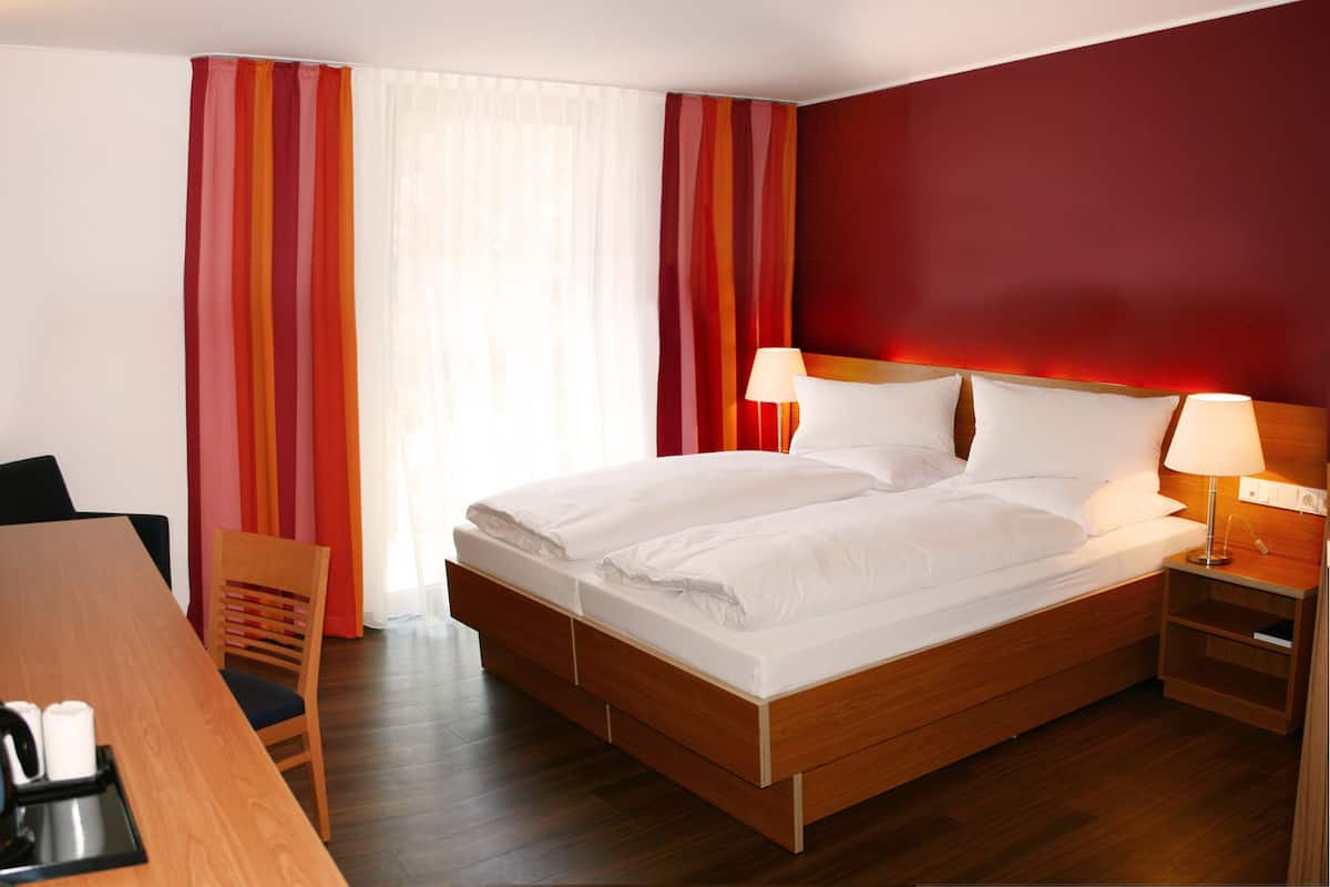 A double bed with white linens, a wooden nightstand with a lamp, a wooden desk with a chair, and a window with sheer curtains.