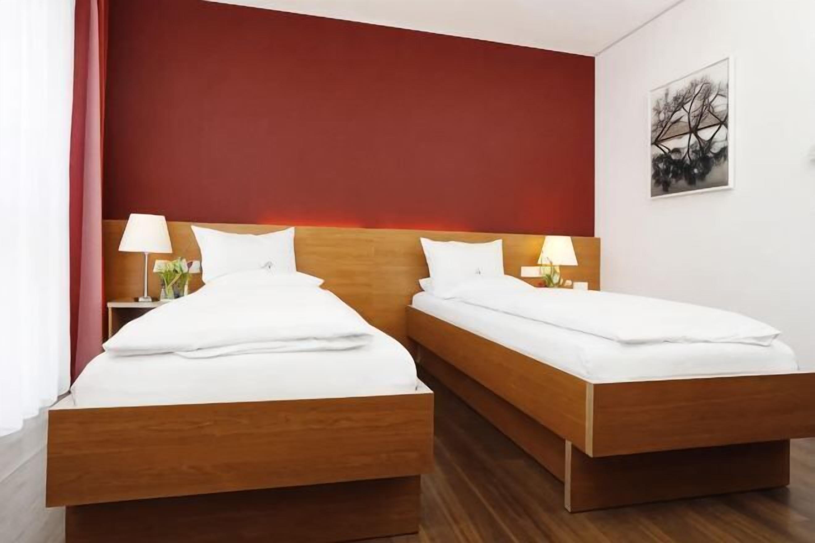 A double room with two beds, each with white bedding and wooden headboards, a red accent wall, and a framed picture on the wall.