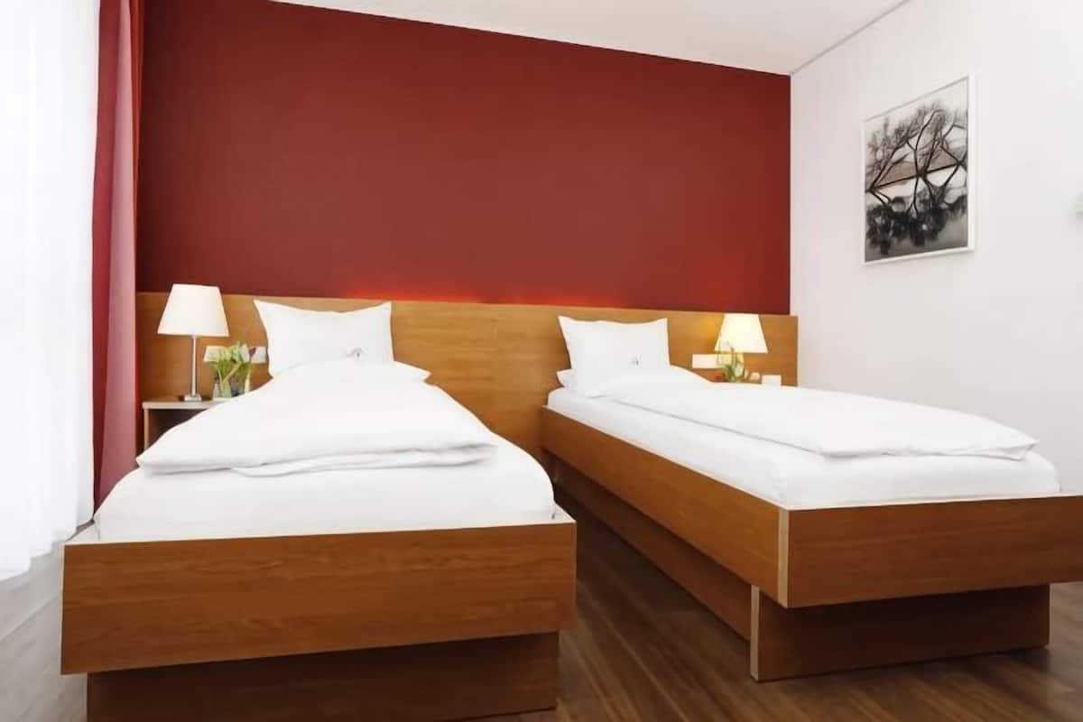 A double room with two beds, each with white bedding and wooden headboards, a red accent wall, and a framed picture on the wall.