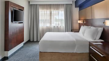Ein Hotelzimmer mit einem großen Bett, einem wandmontierten Flachbildfernseher, einem Nachttisch und einem Fenster mit Vorhängen.
