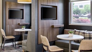 Ein modernes Hotelzimmer mit zwei an der Wand befestigten Fernsehern, einem runden Tisch mit Zimmerpflanze und einer Couch mit karierten Kissen.