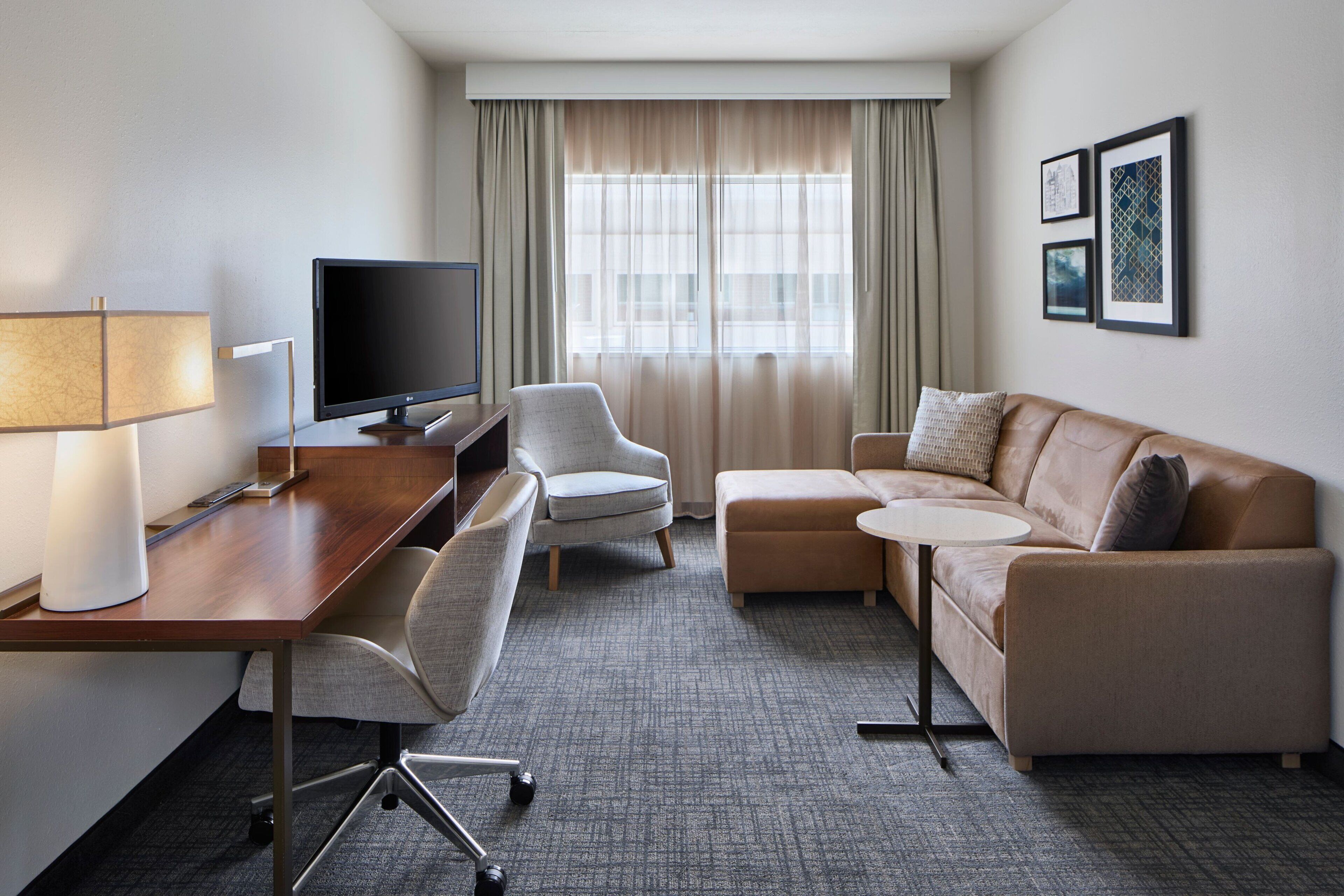 A hotel room with a wooden desk, a flat-screen TV, a grey armchair, a beige sofa, and a small round table.