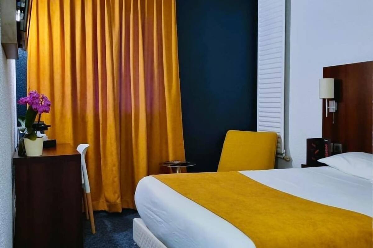 A hotel room with a bed, a desk, a chair, a small table, and a curtain.
