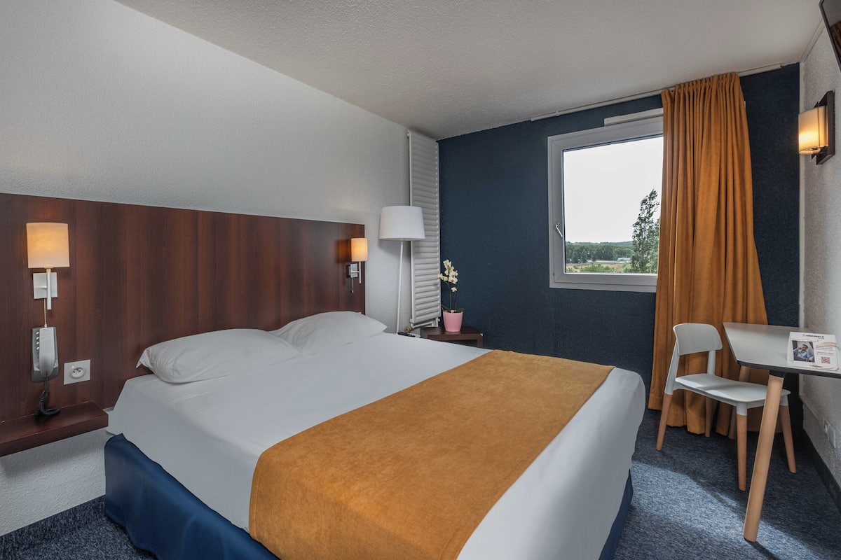 A hotel room with a bed, bedside lamps, a desk with a chair, a window with curtains, and a view of the outdoors.
