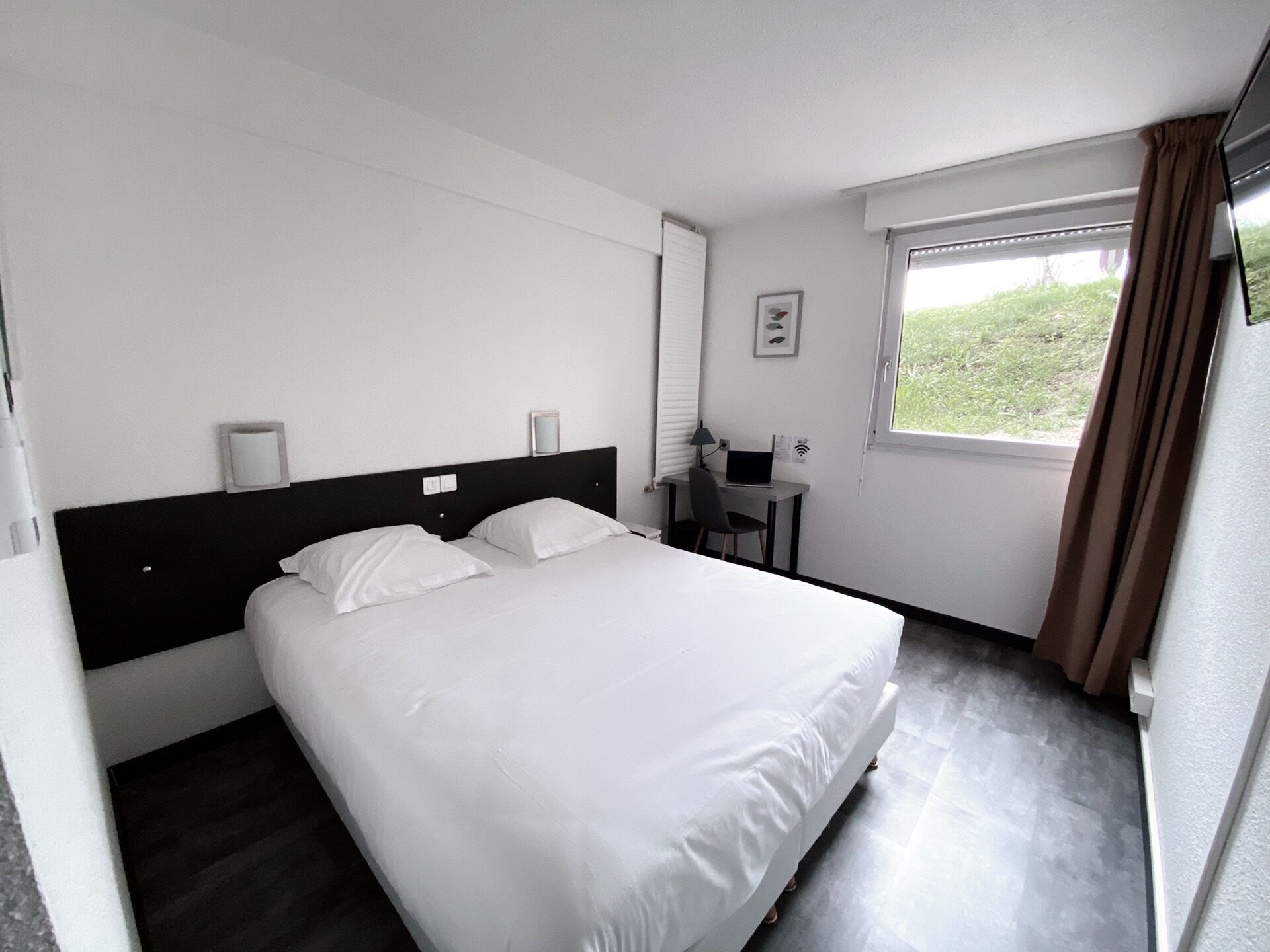A hotel room with a bed, a desk with a laptop, a window with curtains, and a wall-mounted light.