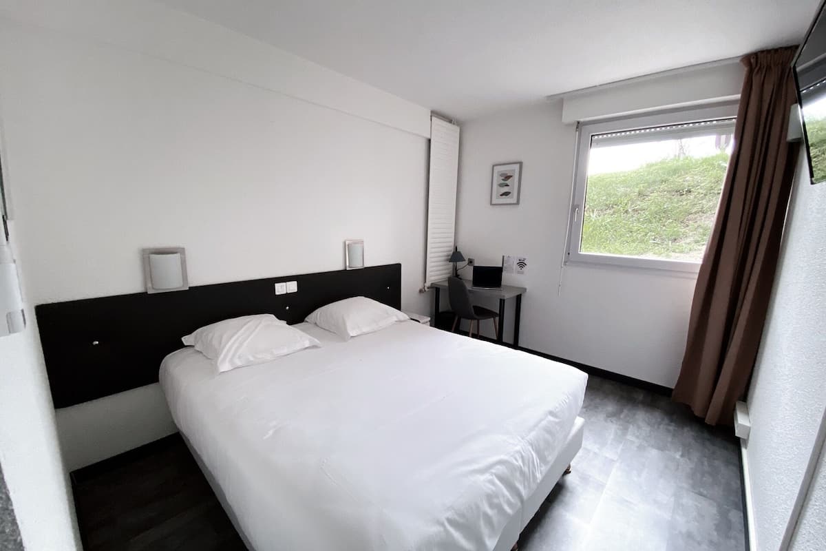 A hotel room with a bed, a desk with a laptop, a window with curtains, and a wall-mounted light.