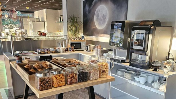 A buffet area with various food items, a coffee machine, and a large moon-themed wall. - The originals Hotel Ariane, Toulouse (Toulouse)