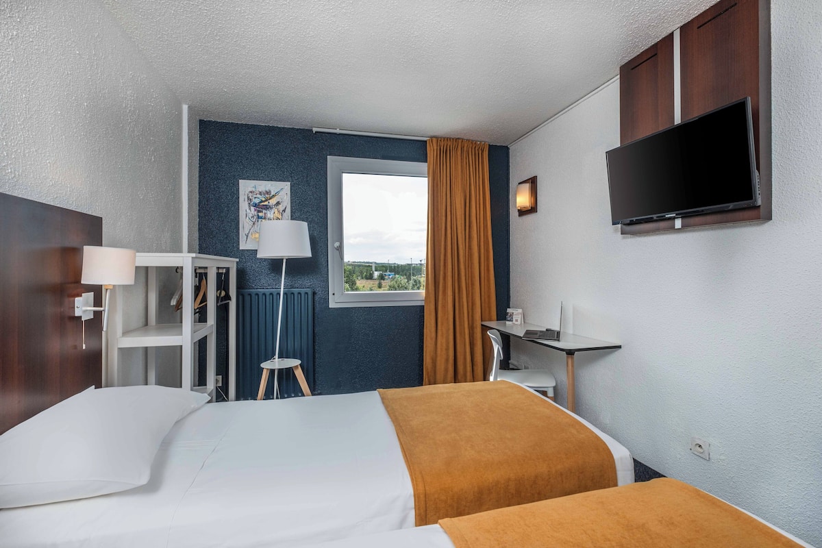 A hotel room with two beds, a desk, a chair, a TV mounted on the wall, a window with curtains, and a radiator.